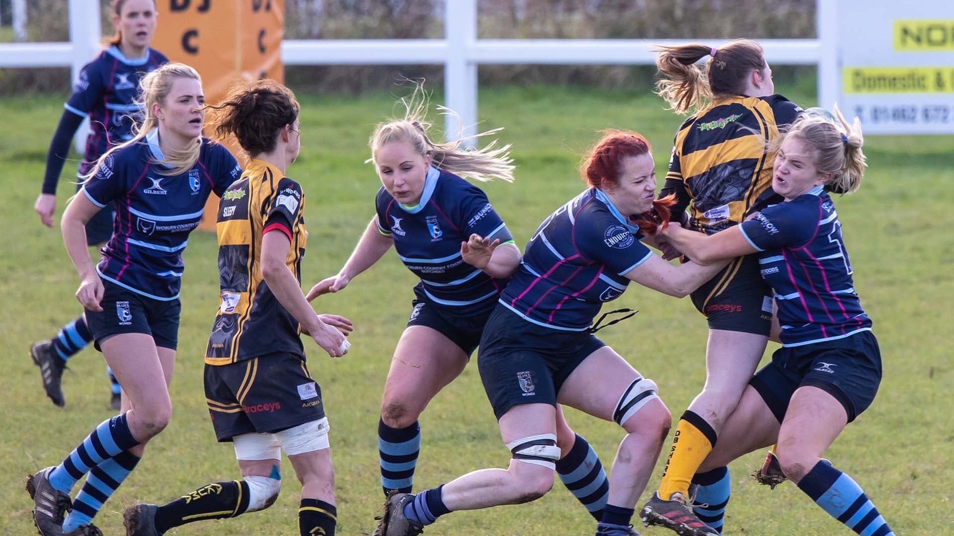 An adult women's match, a player in navy and sky blue driving into a tackle with team-mates in support