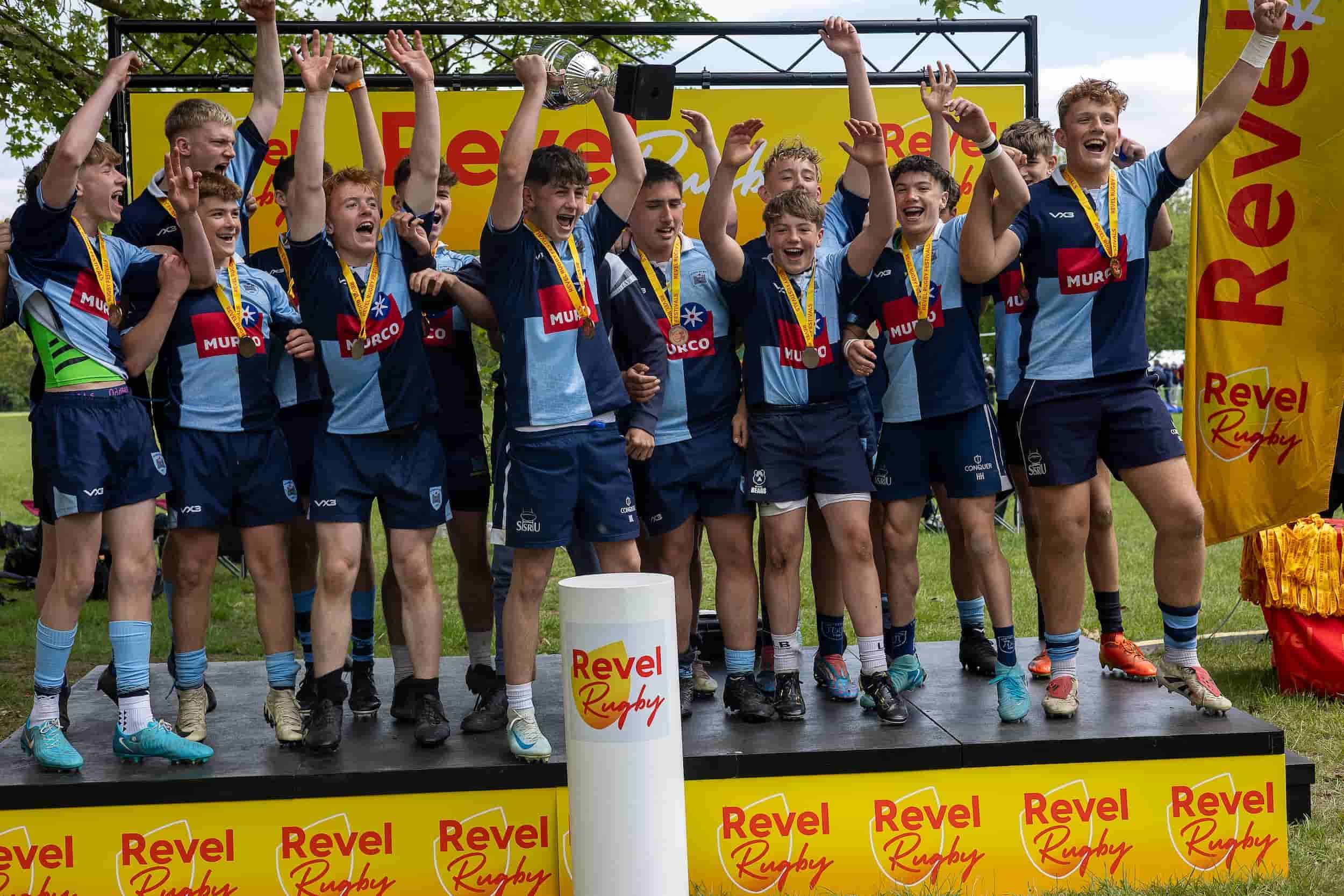 A senior-age junior squad in sky and navy celebrating on the stage with medals and the trophy