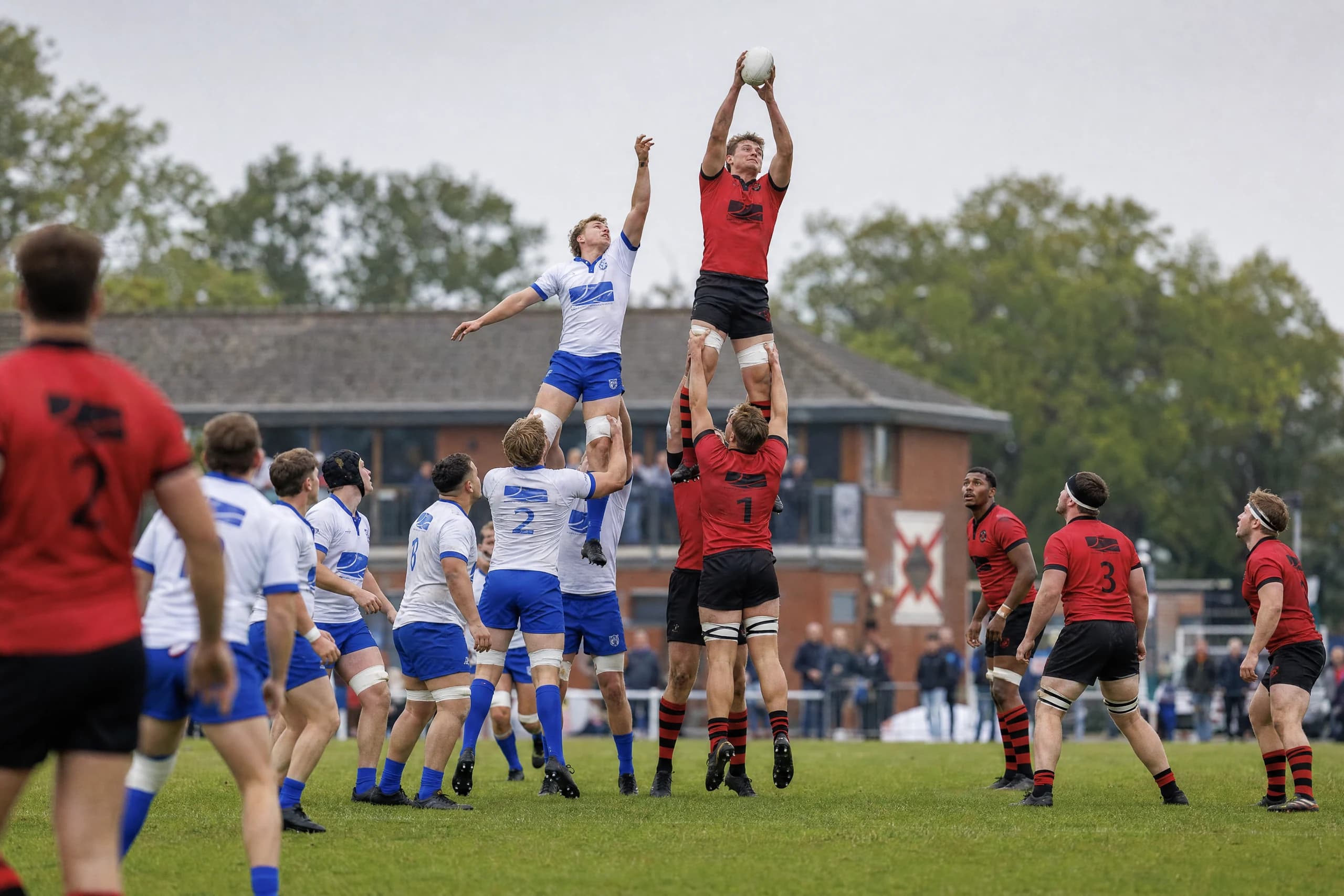 A lineout jumper in red and black at full stretch to catch the ball, with the clubhouse behind